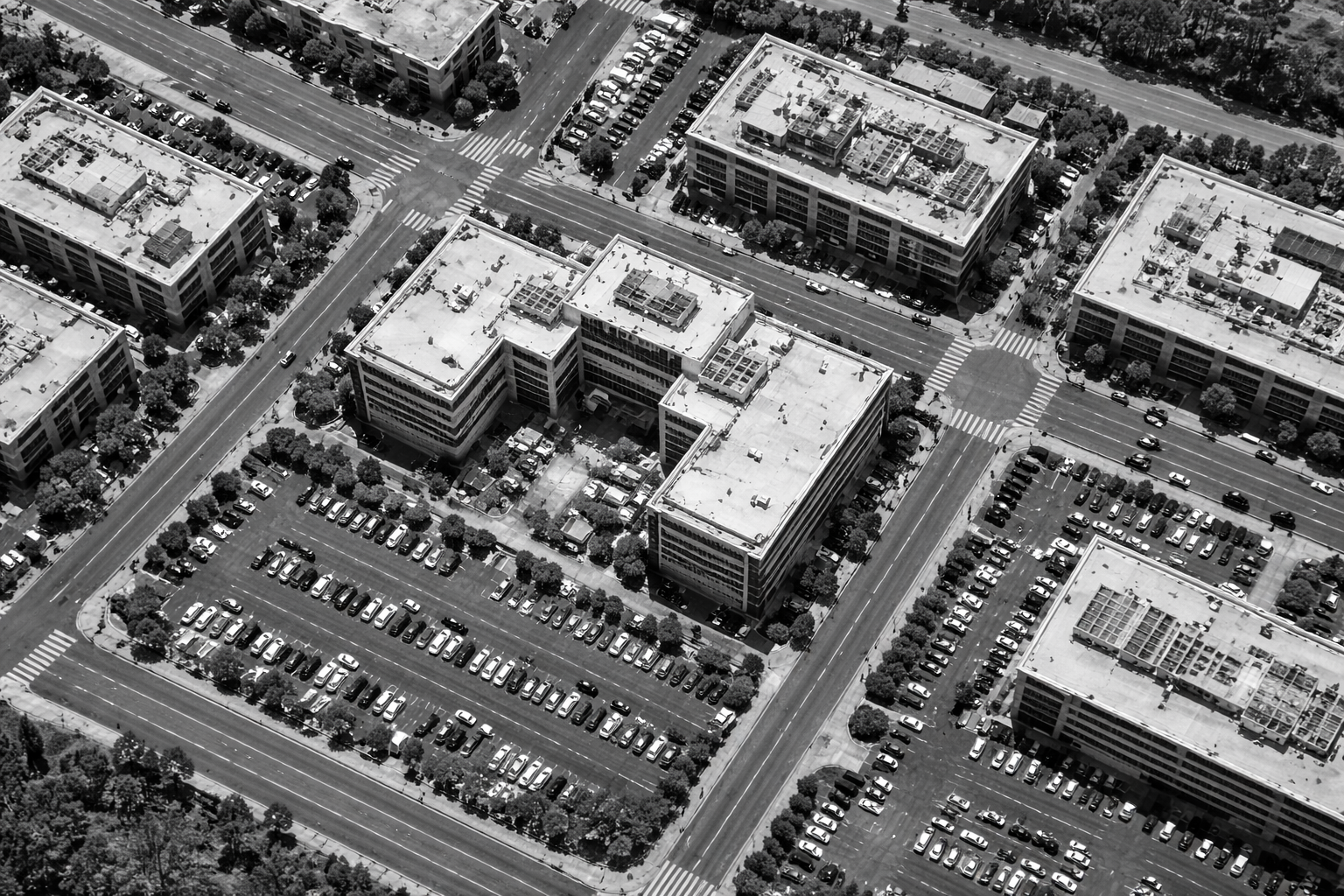 Black and white aerial view of a dense city block with intersecting streets, rooftops, parked cars, and surrounding commercial buildings.